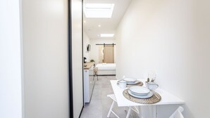 Une chambre d’hôtel moderne et minimaliste, aux couleurs claires, avec un lit doté d’une tête de lit en bois, un petit bureau avec une lampe et un coin repas avec une table dressée pour deux personnes.