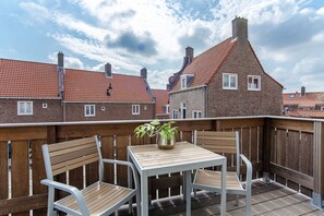 A wooden deck with a table and two chairs, a potted plant, and a view of red-tiled buildings under a partly cloudy sky. - De Boulevard 2 D (Zandvoort)