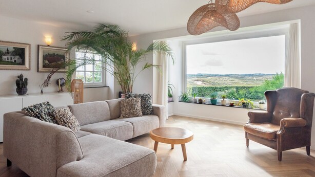 A living room with a sofa, armchair, and round wooden table. There is a large window with a view of greenery and hills. - Family villa behind the Beach. For 3 couples over 30 years old. (Zandvoort)