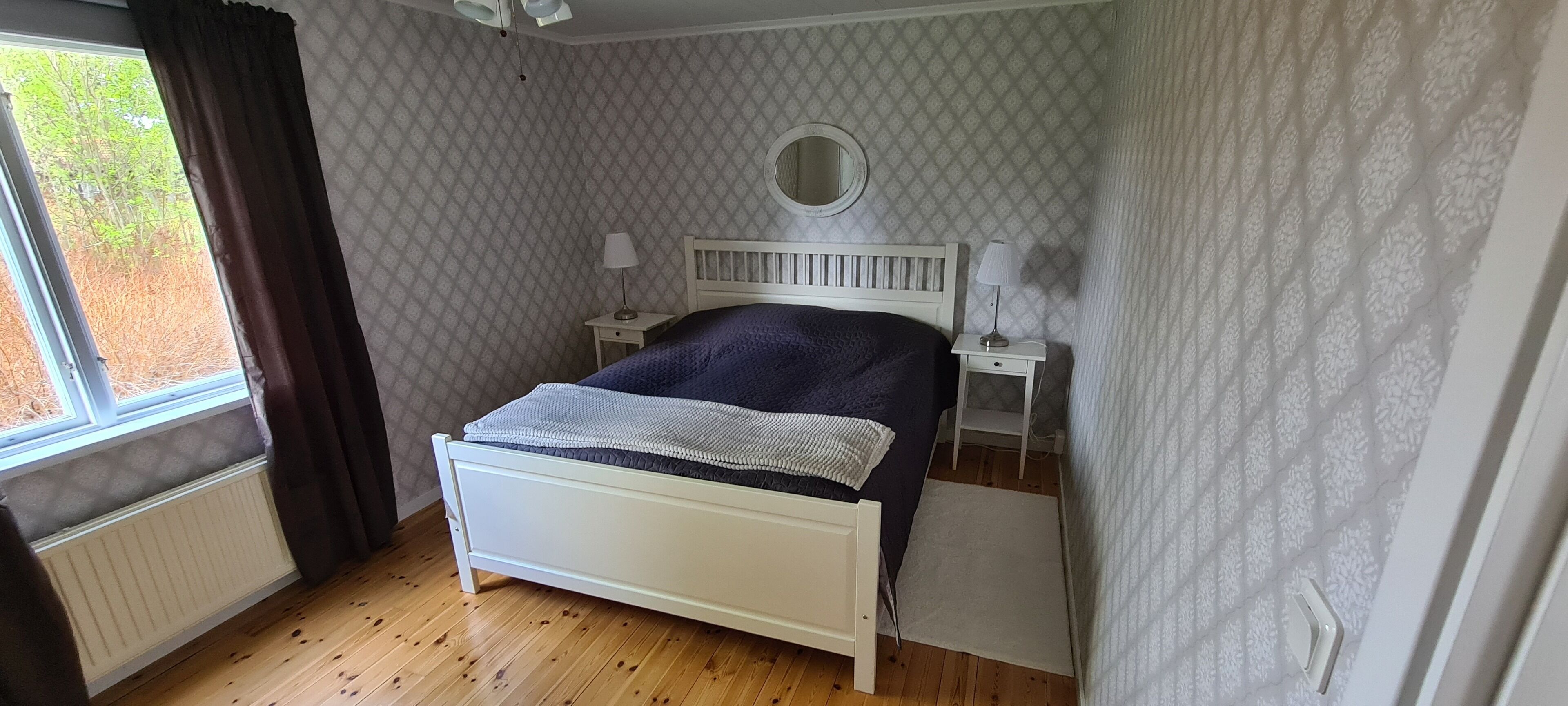 A bedroom with a white bed, a window with curtains, a bedside table with a lamp, and a patterned wall.