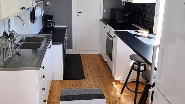 A compact kitchen with white cabinets, a black stove, and a wooden floor. - Hel Stuga för 4 i Härlig Bymiljö (Dalarnas län)