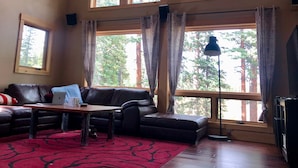 Una sala de estar con un sofá de cuero, una mesa de centro de madera y un tapete rojo estampado. Hay ventanas amplias con cortinas, una lámpara de pie y un televisor.