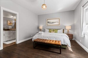 A bedroom with a bed, bedside lamps, a bench, a framed picture on the wall, and a bathroom visible through an open door. - Spacious Family Home with Hot Tub and Game Room (Coeur d'Alene)
