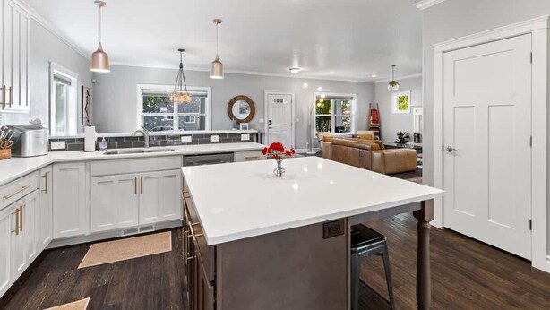 A modern kitchen with white cabinetry, a central island, pendant lighting, and a dining area with a sofa. - Family fun home with hot tub, arcade and zipline (Coeur d'Alene)