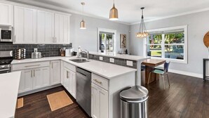 A modern kitchen with white cabinets, a stainless steel dishwasher, and a wooden dining table. - Family fun home with hot tub, arcade and zipline (Coeur d'Alene)