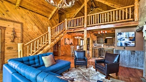 Hirsihirrestä rakennetun mökin sisustus: sininen sohva, nahkaiset nojatuolit, puinen portaikko ja keittiötila.