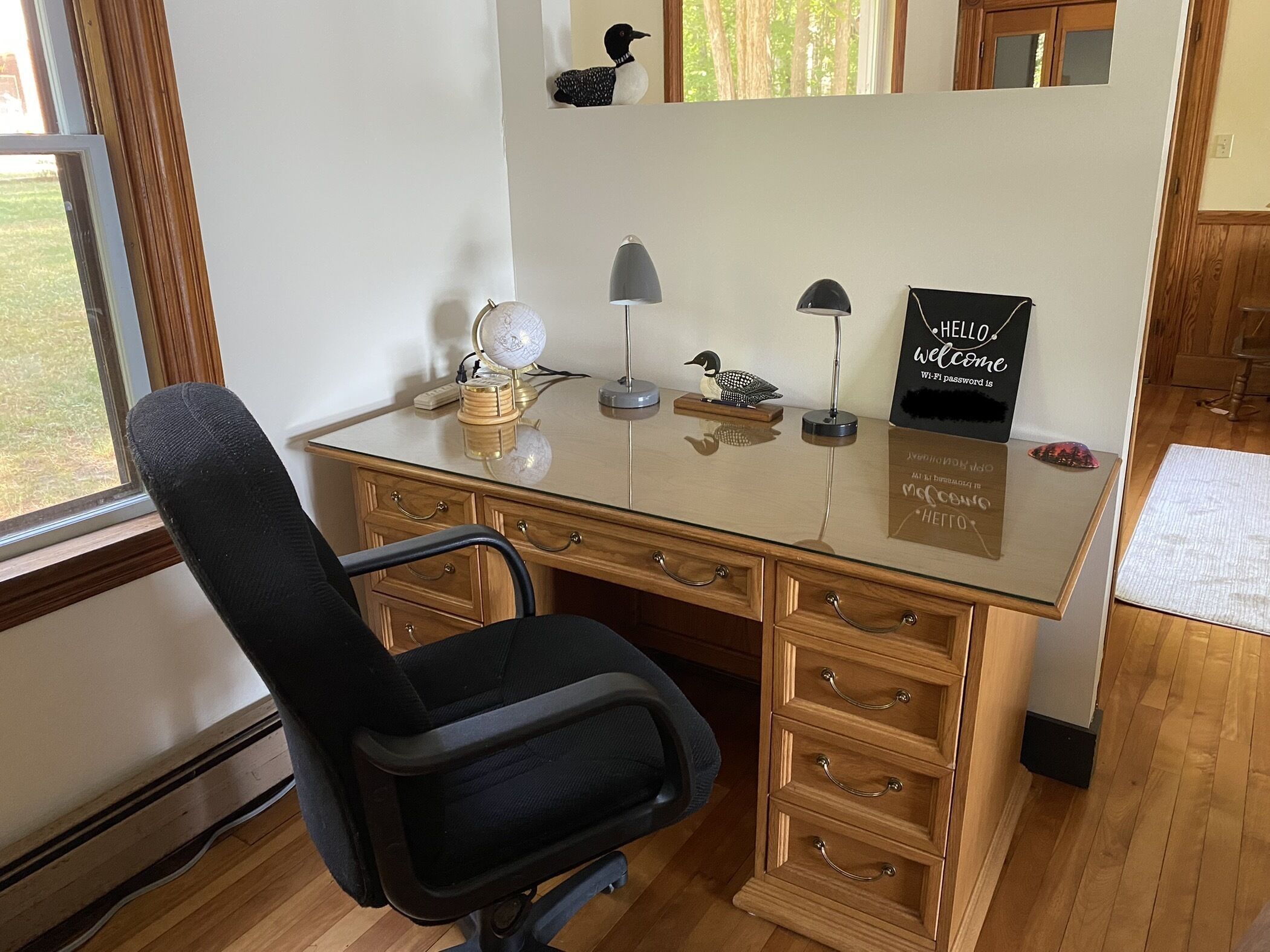 Un escritorio de madera con cubierta de vidrio, una silla de oficina negra, un globo terráqueo, una lámpara y un cartel con la contraseña de Wi-Fi.