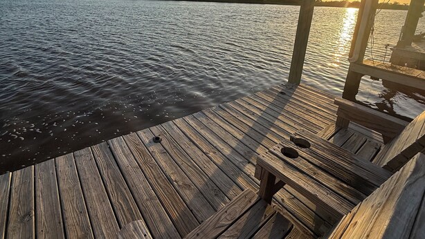 A wooden dock extending over calm water with a clear sky and a sunset in the background.