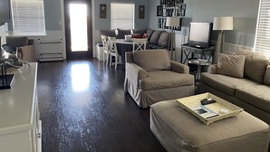 A living room with a dark wooden floor, beige sofas, a dining area with a white table and chairs, and a ceiling fan.
