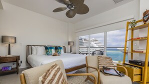 Een hotelkamer met een bed, twee fauteuils, een houten bijzettafeltje, een boekenkast en een groot raam met uitzicht op een cruiseschip.