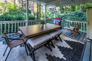 A covered outdoor patio with a wooden table and chairs, a small grill, and a view of a lush garden.