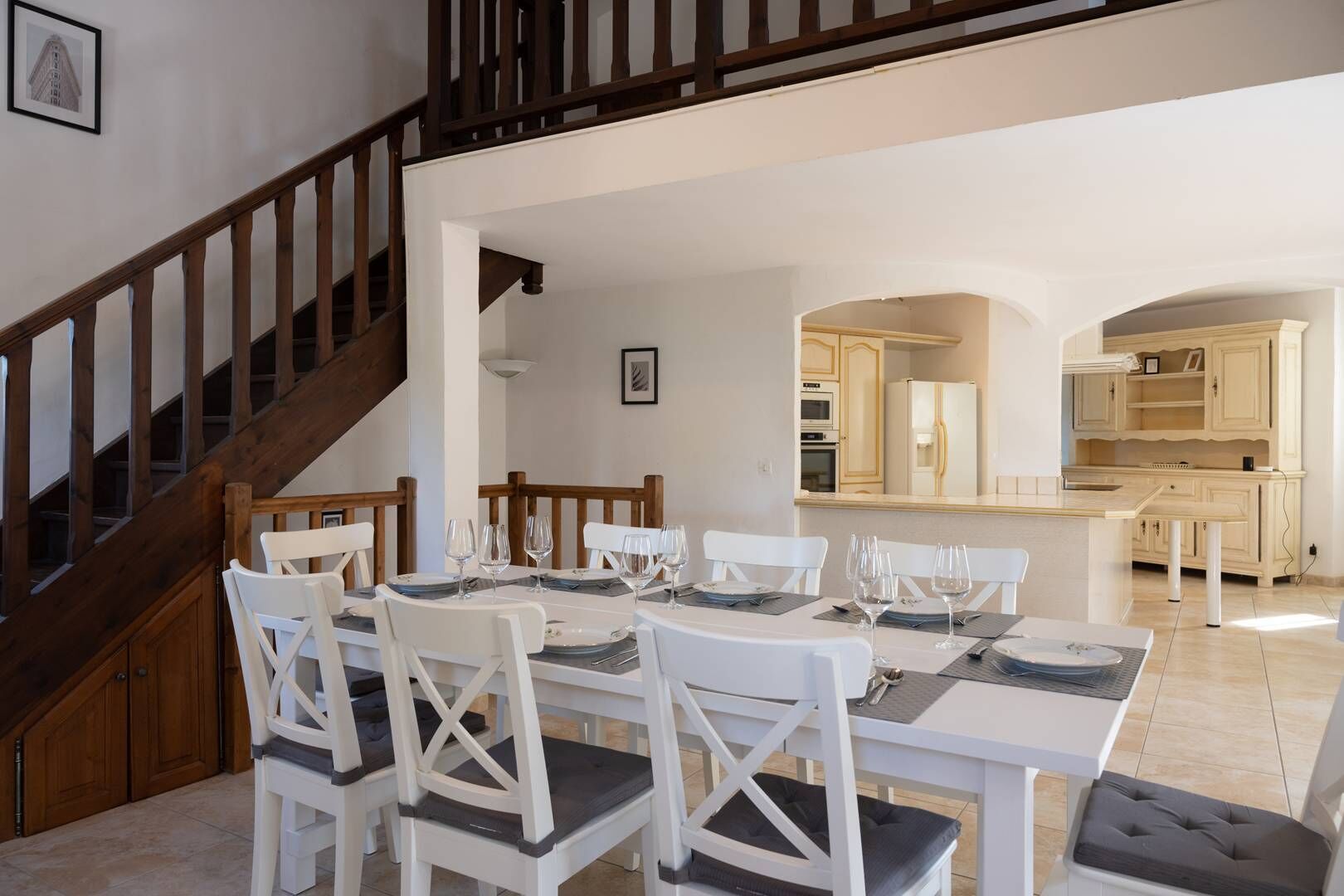 Un área de comedor con una escalera de madera, sillas blancas y una cocina al fondo.