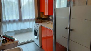 Un espace réduit dédié à la buanderie, comprenant un lave-linge, un évier et une petite fenêtre avec des rideaux.