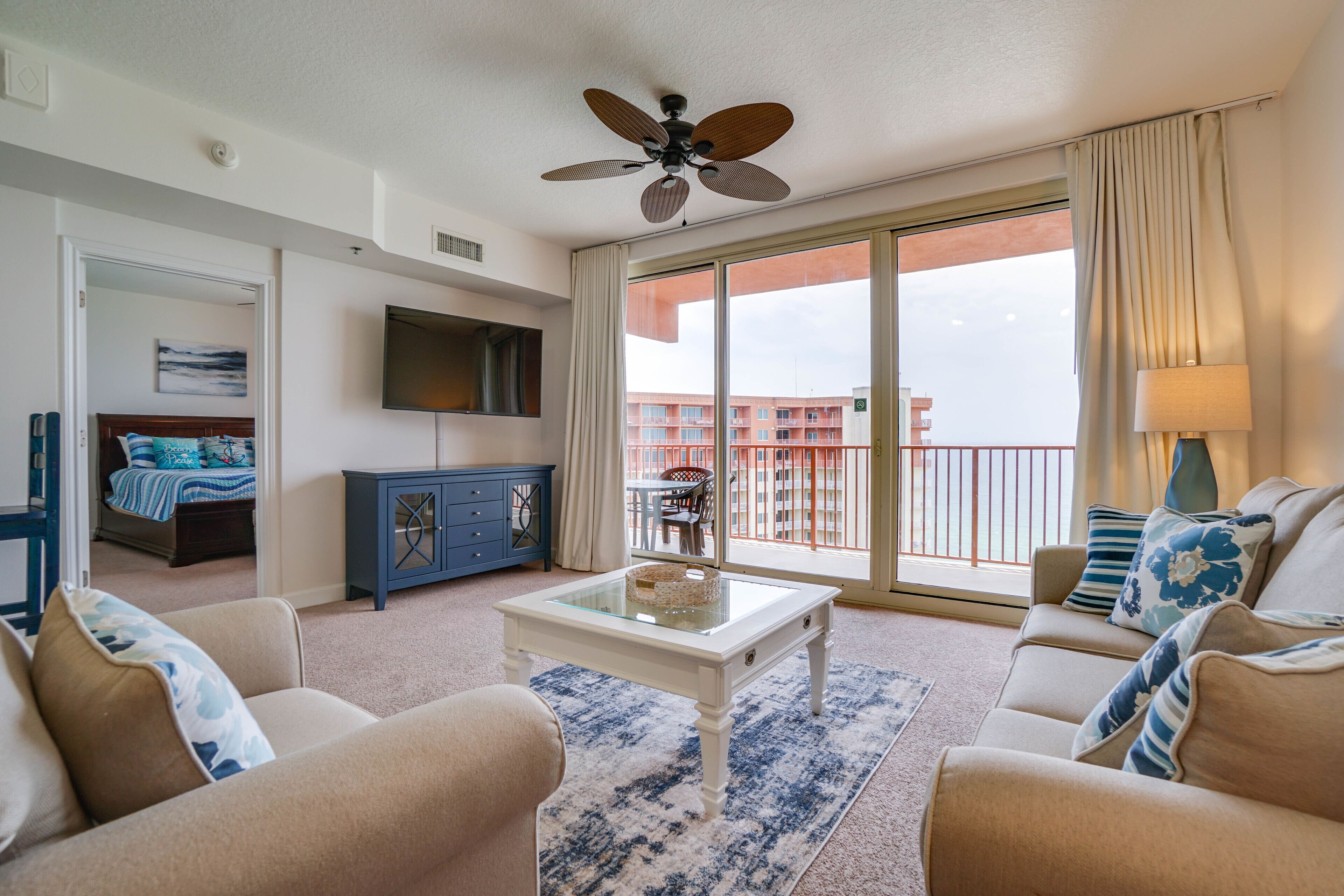 A hotel room with a large balcony, a ceiling fan, a flat-screen TV, a sofa, a coffee table, and a view of the cityscape.