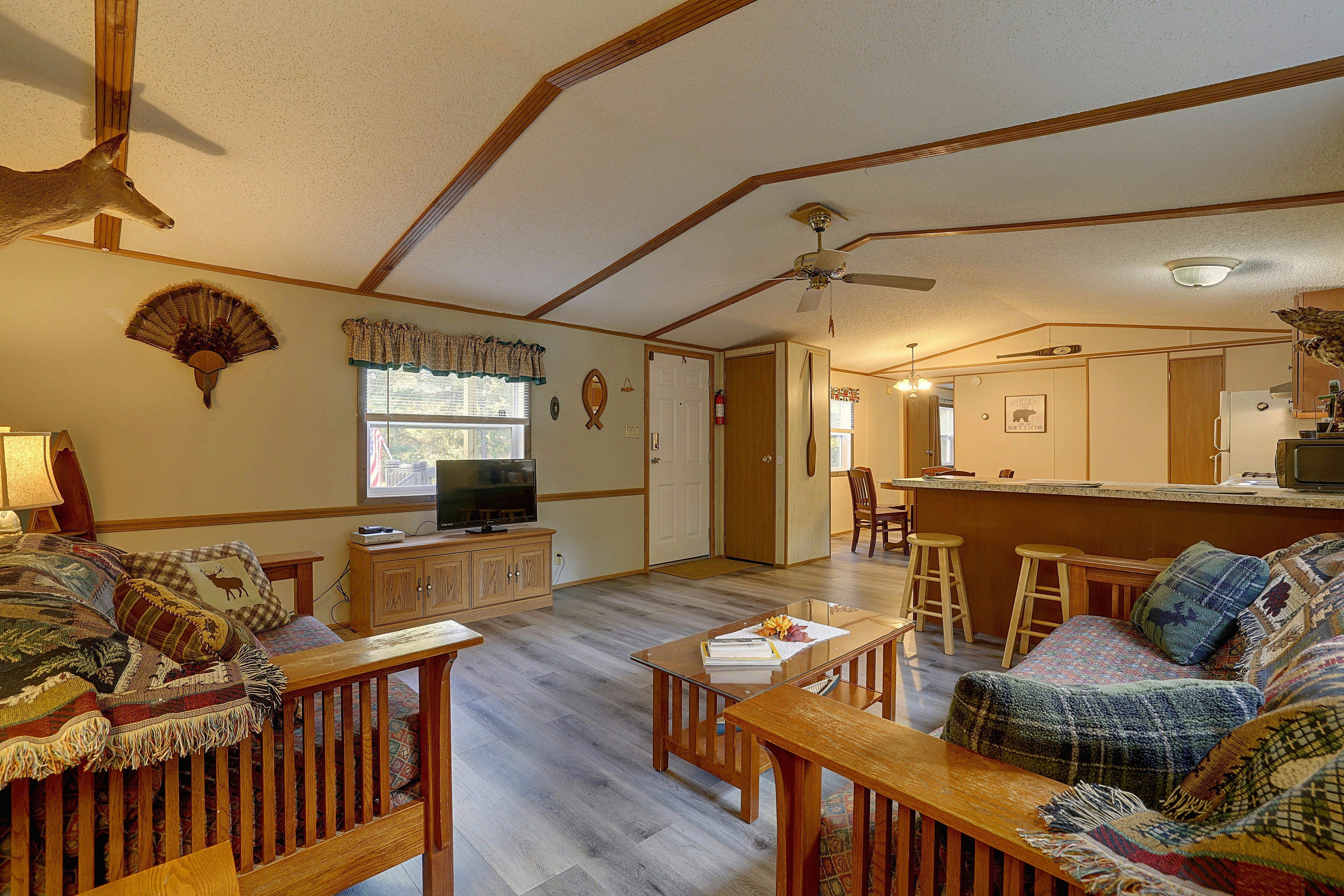 A living room with a television, wooden furniture, a ceiling fan, and a kitchen area with a refrigerator and microwave.