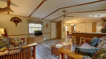 A living room with a television, wooden furniture, a ceiling fan, and a kitchen area with a refrigerator and microwave.