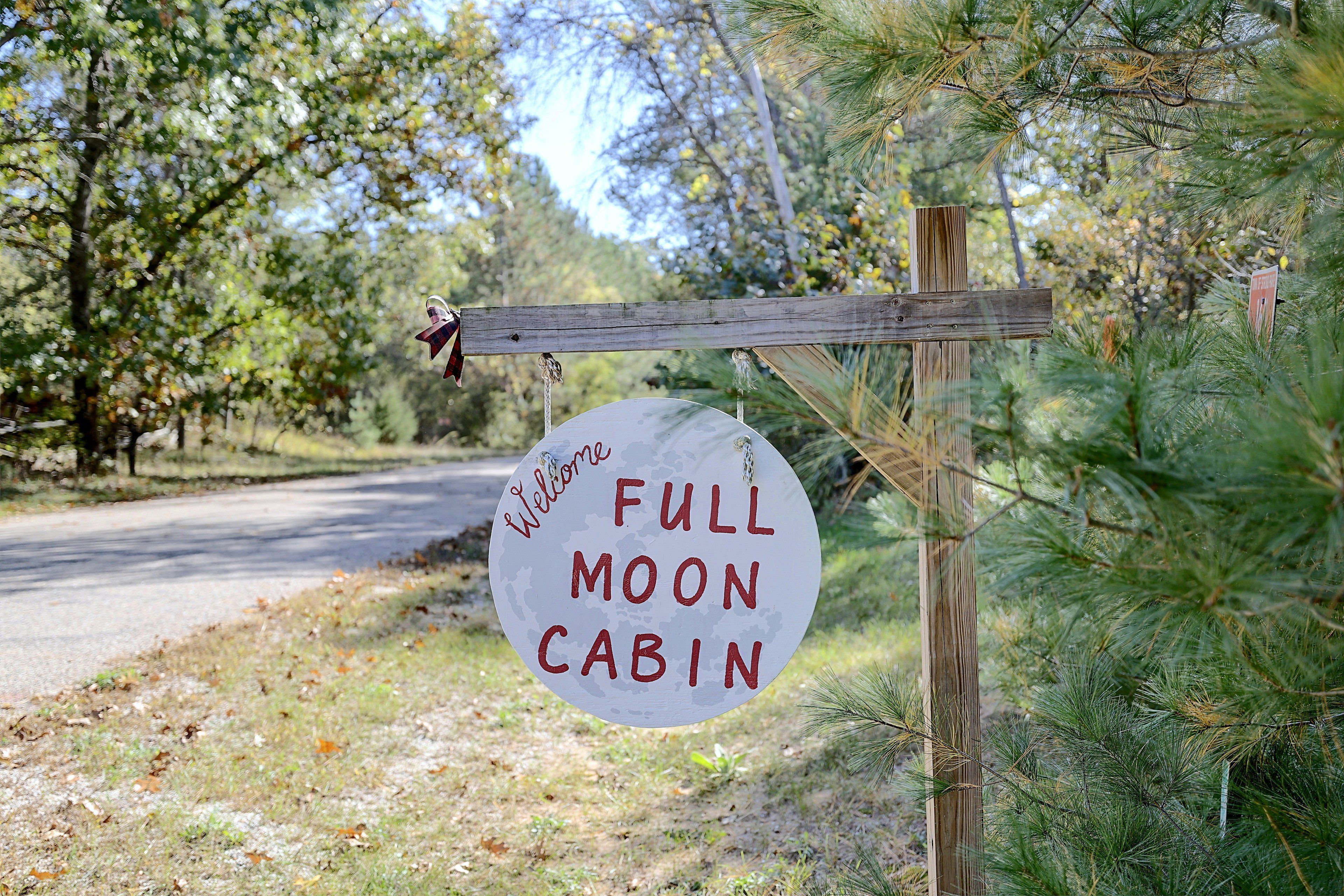 A wooden sign with 'Welcome Full Moon Cabin' written on it, hanging on a post with a wooden fence.