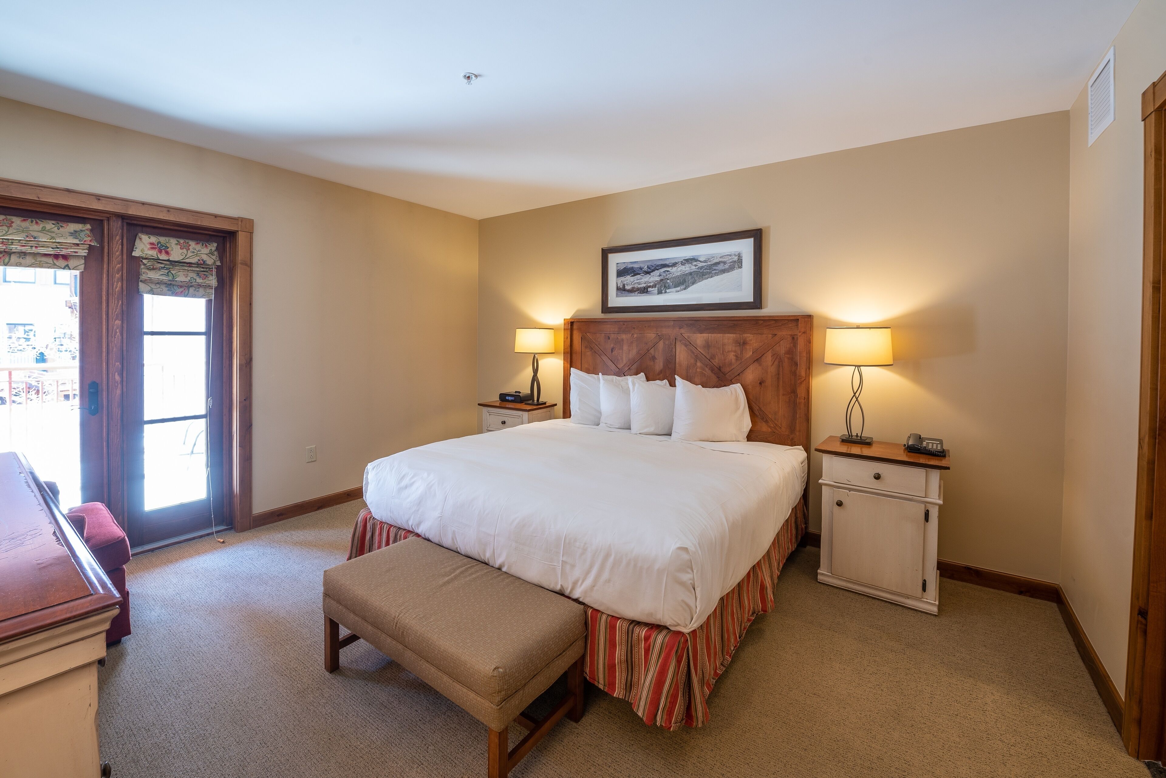 A neatly arranged bedroom with a large bed, bedside tables with lamps, a bench, and a door leading to an outdoor area.