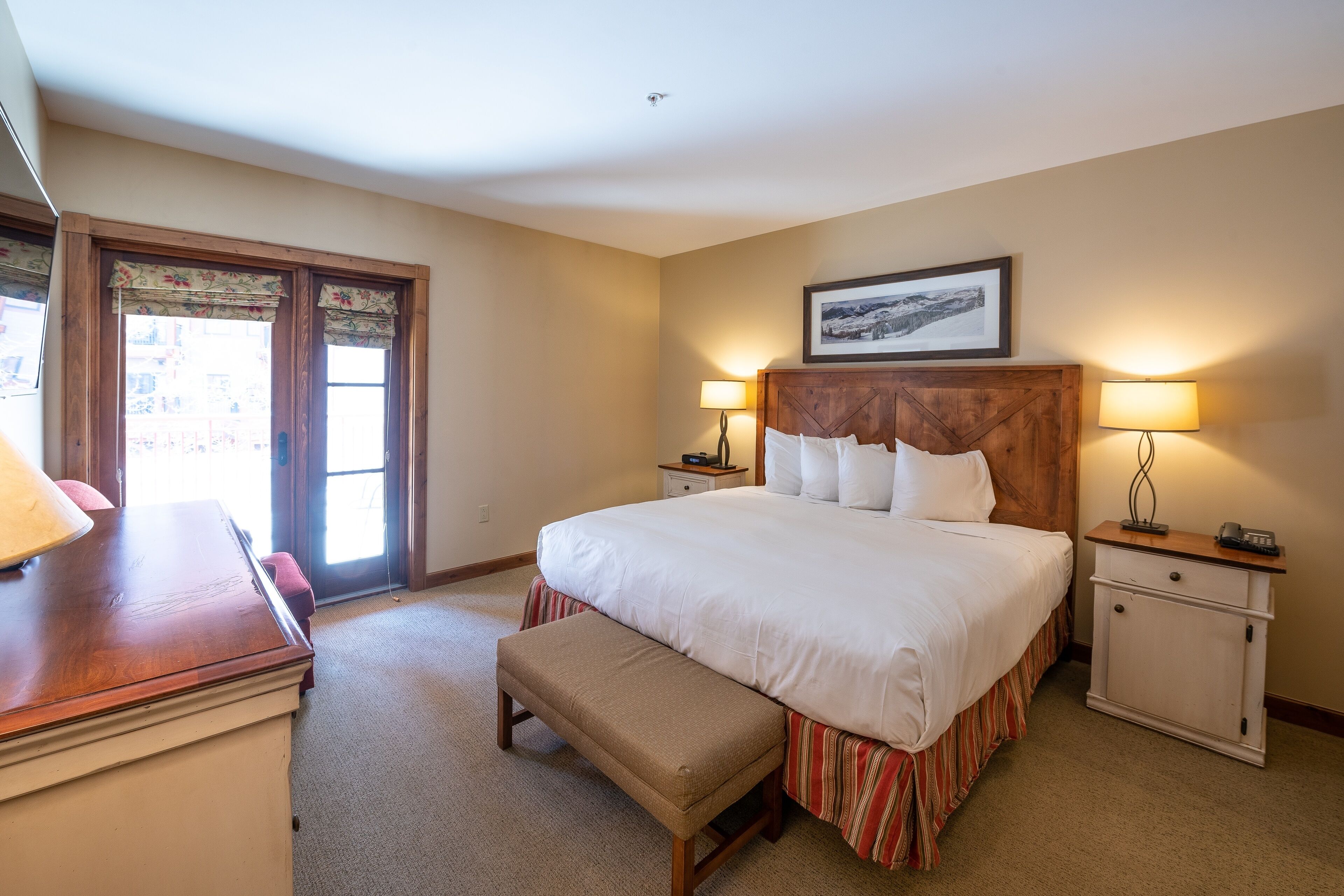 A hotel room with a large bed, two bedside tables with lamps, a bench, and a desk with a lamp.