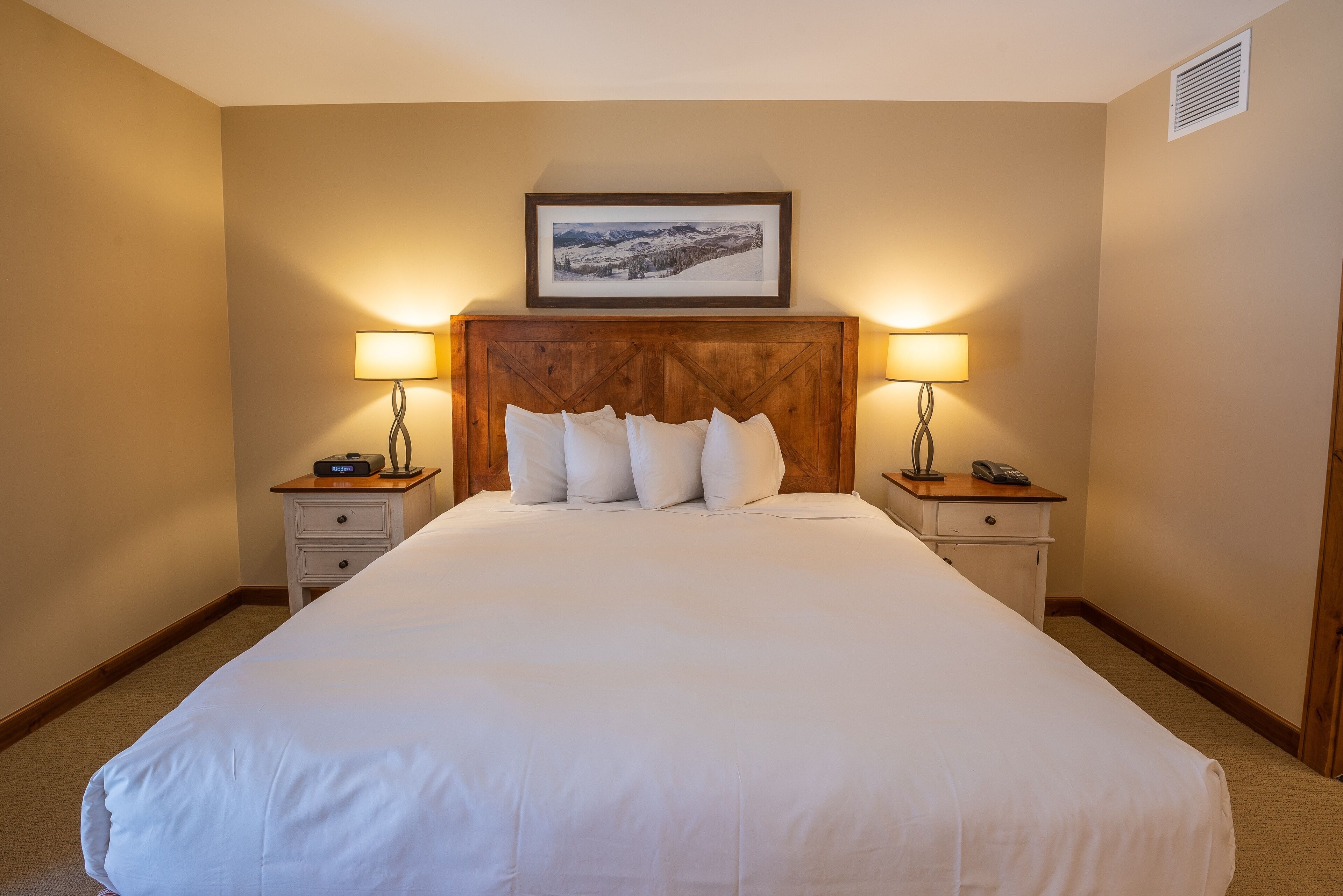 Habitación de hotel con una cama grande, dos mesitas de noche con lámparas, un cuadro enmarcado en la cabecera y un cajón pequeño.