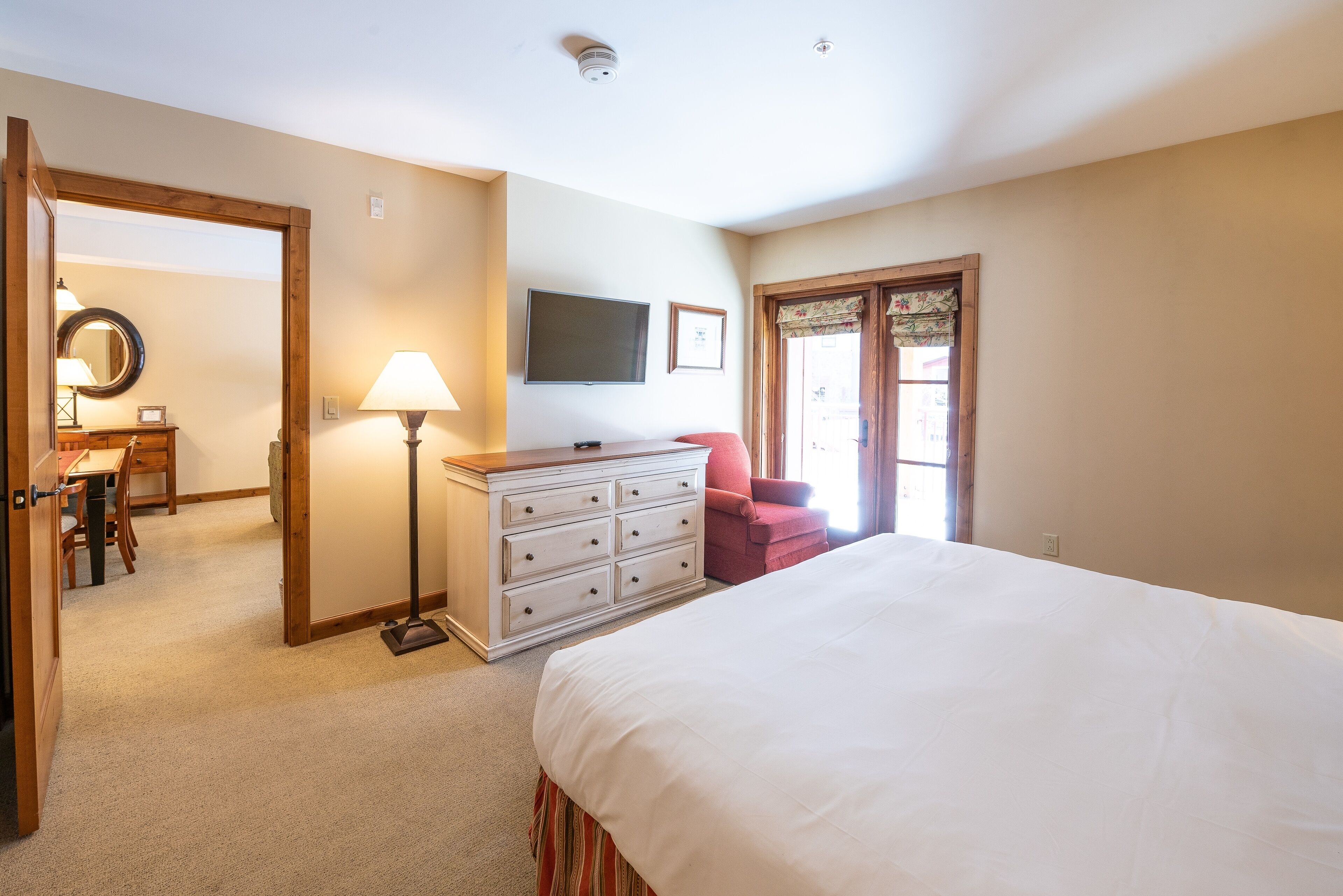 A bedroom with a bed, dresser, desk, chair, lamp, TV, and a door leading to another room.