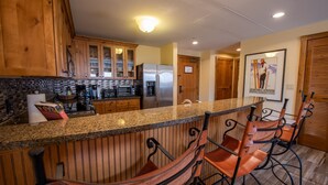 A kitchen with wooden cabinets, a granite countertop, a stainless steel refrigerator, and a bar area with two chairs.