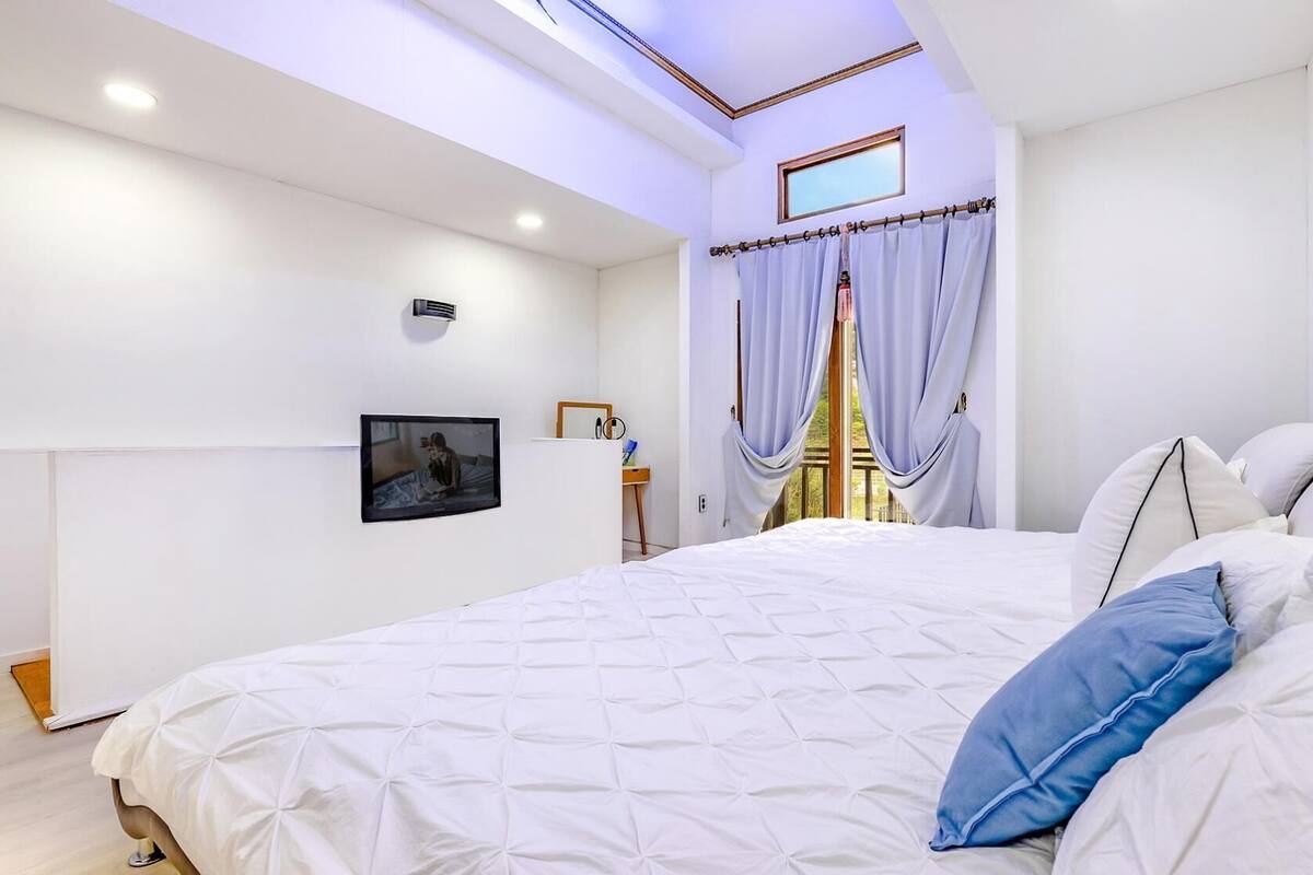 A bedroom with a bed, a TV mounted on the wall, and a window with curtains.