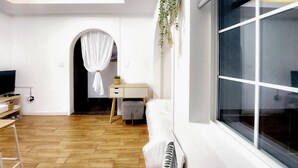 Un salon moderne avec un plancher en bois, un canapé blanc, une petite table sur laquelle est posée une plante, et une grande fenêtre.