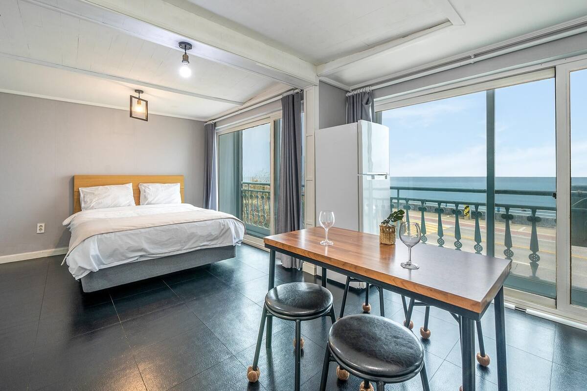 Ett hotellrum med en säng, ett bord med två stolar, utsikt över havet genom stora fönster och ett balkongräcke.