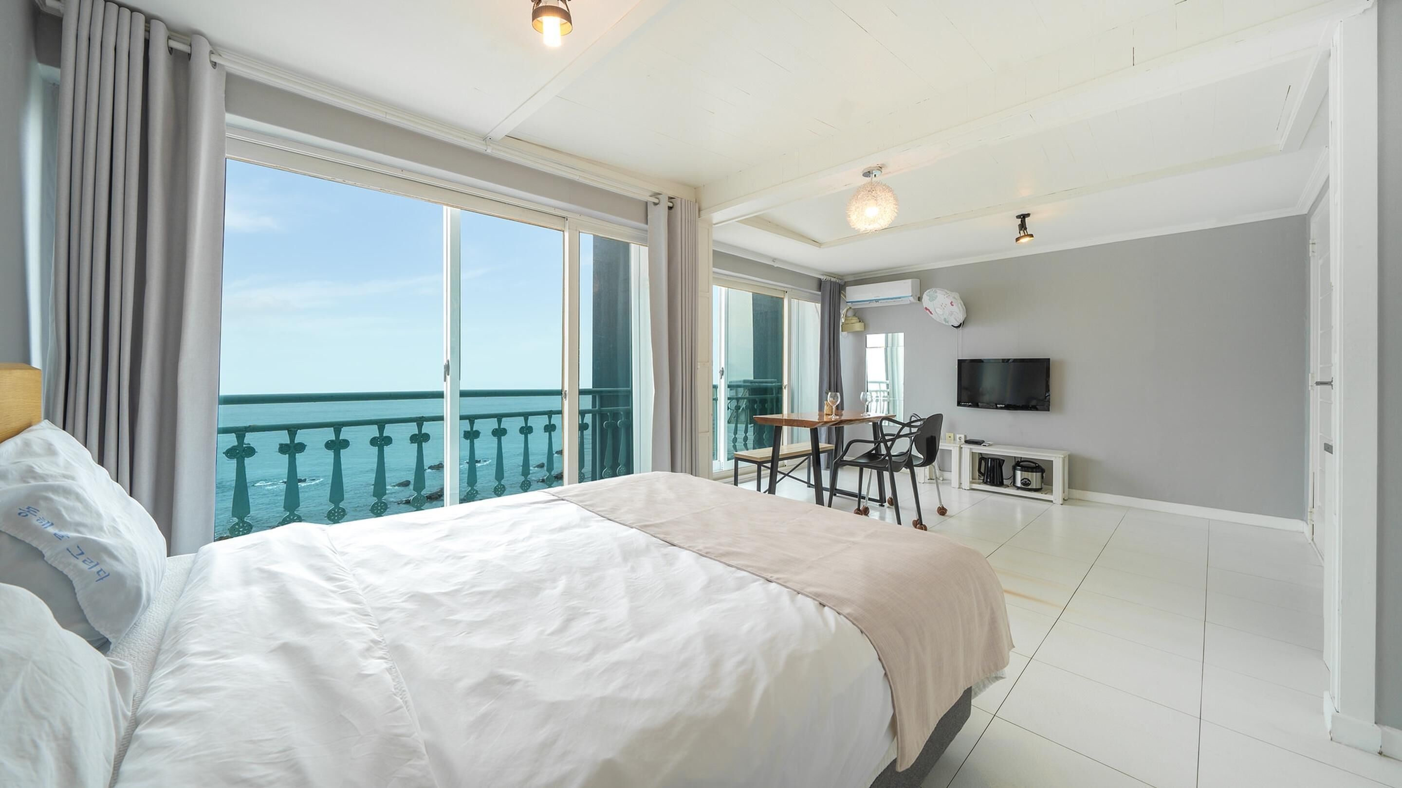 A hotel room with a large bed, a balcony with a view of the sea, a dining table, and a TV.