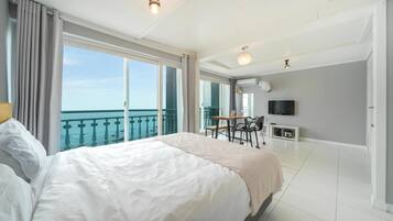 A hotel room with a large bed, a balcony with a view of the sea, a dining table, and a TV.