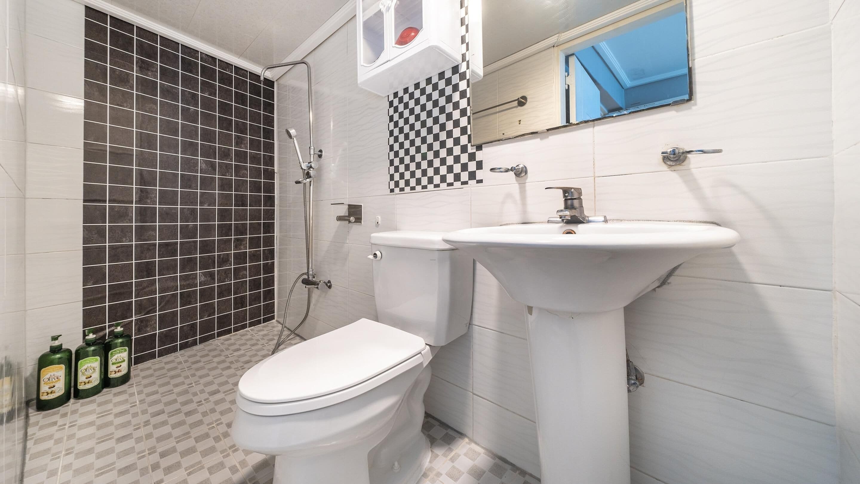 A bathroom with a white toilet, a white sink, and a shower. There are three bottles of olive oil on the floor.