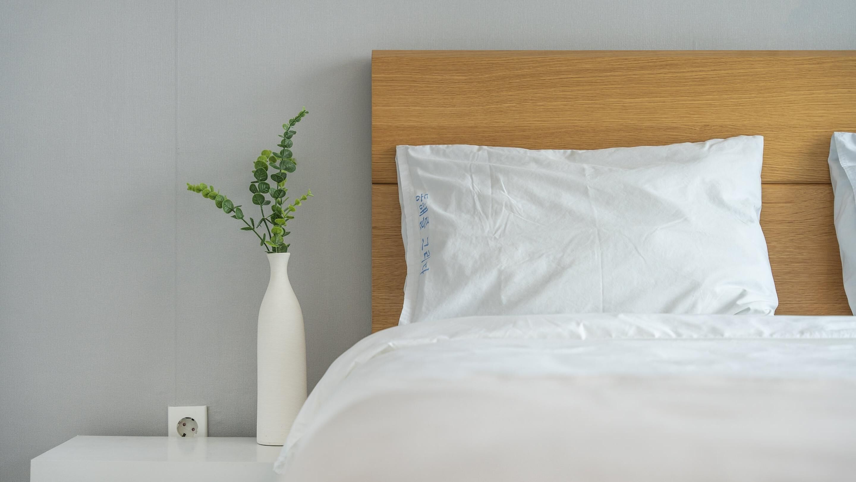 A neatly made bed with white bedding, a wooden headboard, and a white vase with green foliage on a bedside table.