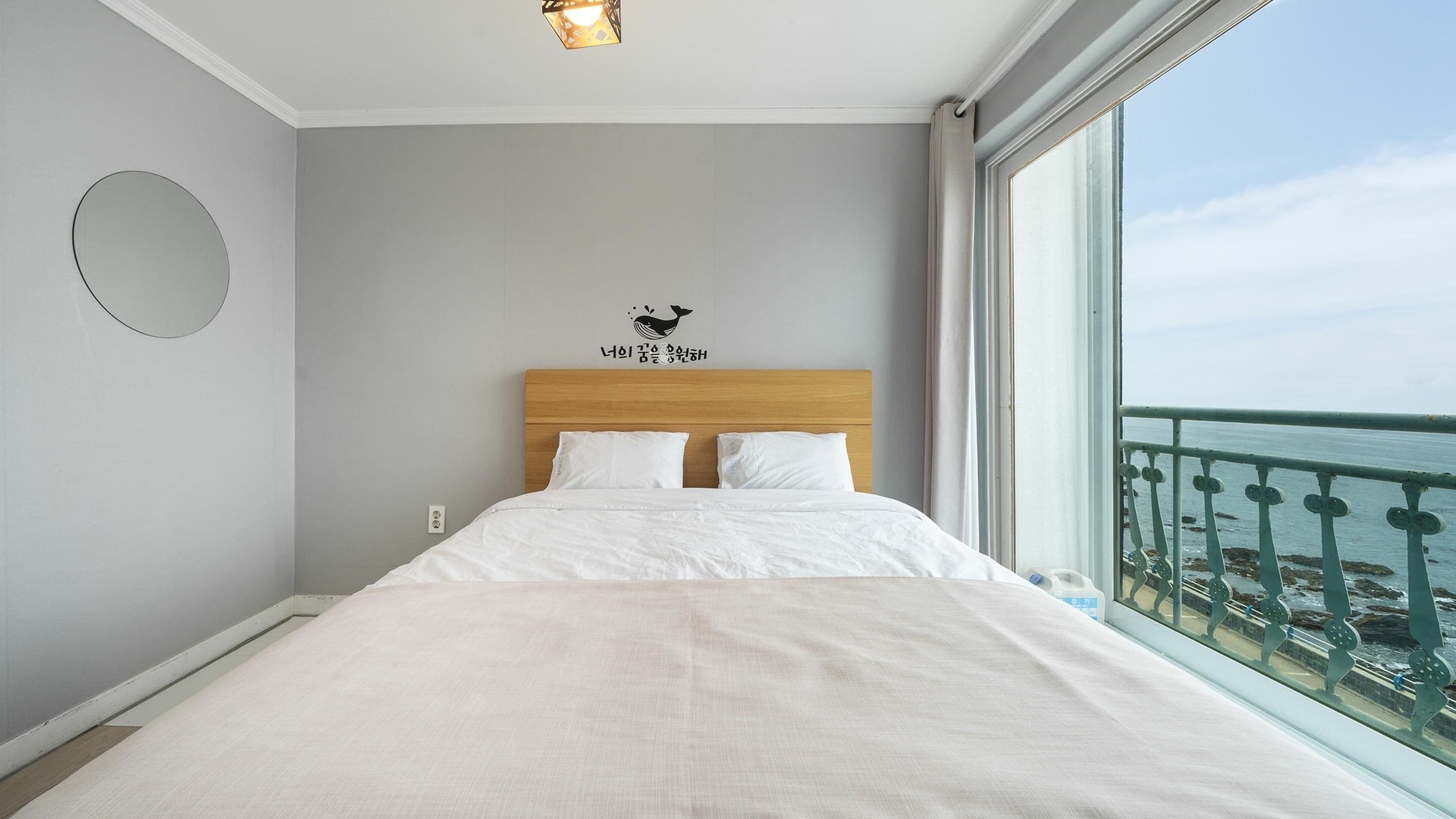 A hotel room with a large bed, a wooden headboard, a wall-mounted circular mirror, and a window with a view of the sea.