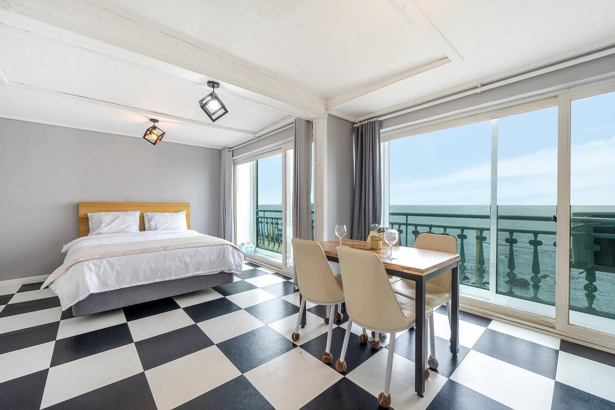 Ett hotellrum med ett rutmönstrat golv, en säng med vita sängkläder, ett matbord med stolar och stora fönster med utsikt över havet.