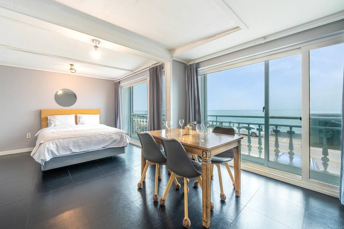 Ett hotellrum med en säng, ett matbord med stolar, ett stort fönster med utsikt över havet och en balkong.