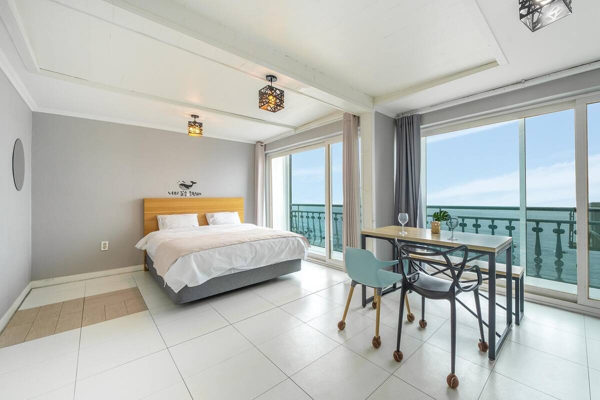 Ett modernt hotellrum med en säng, ett matbord med stolar, en balkong med utsikt över havet och stora fönster.