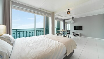 Habitación de hotel con una cama grande, zona de comedor con mesa y sillas, televisor montado en la pared y balcón con vista al mar.