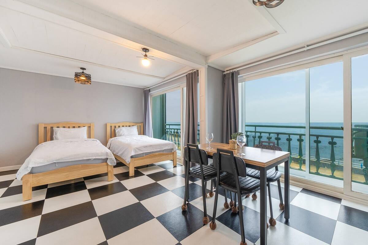 Ett hotellrum med två sängar i trä, ett rutmönstrat golv, ett matbord med stolar och stora fönster med utsikt över havet.