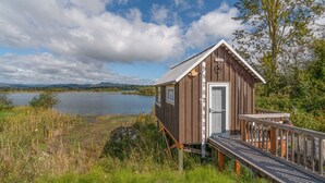 A wooden cabin with a small deck and a door, situated by a lake with a clear sky. - Dog Friendly Puget Island Home on the Columbia River. Secure Private Moorage!! (Cathlamet)