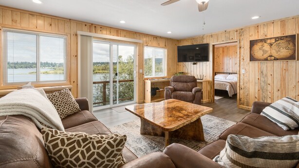 A cozy cabin interior with a wooden exterior, a large window with a view, a ceiling fan, and a flat-screen TV mounted on the wall. - Dog Friendly Puget Island Home on the Columbia River. Secure Private Moorage!! (Cathlamet)