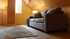 A cozy room with a wooden ceiling, a grey sofa with patterned cushions, a blue patterned rug, and a floor lamp.