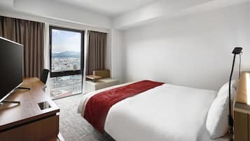 A hotel room with a bed, a desk, a chair, a TV, and a view of a cityscape through large windows.