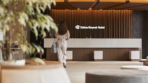 Un área de recepción con diseño moderno, que incluye un mostrador de recepción, un cartel de Dalwa Roynet Hotel y una persona caminando hacia el mostrador.