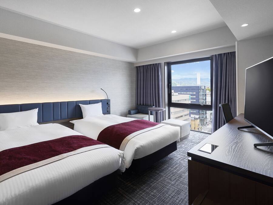 A hotel room with two beds, a desk, and a large window offering a city view.