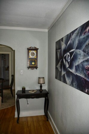 A hallway with a wooden floor, a wall-mounted clock, a table with a lamp, and a large abstract painting.