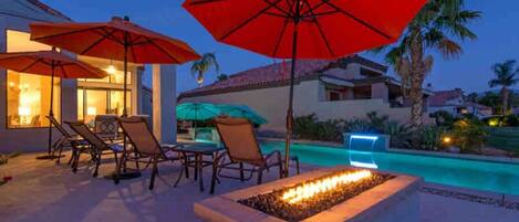 A poolside area with lounge chairs and umbrellas, a fire feature, and a view of a residential building.