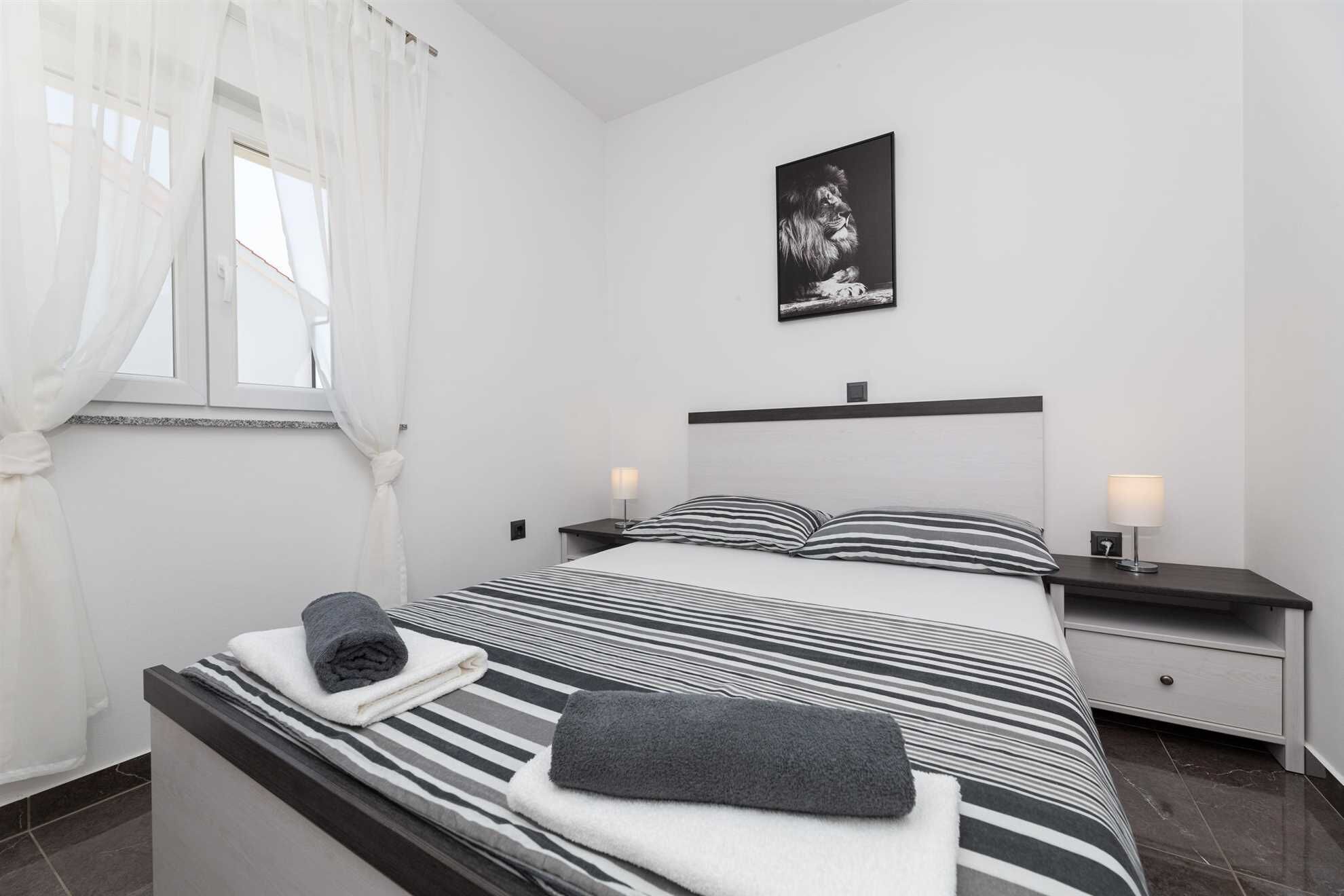 A modern bedroom with a bed, two bedside tables, a nightstand, a window with sheer curtains, and a framed picture on the wall.