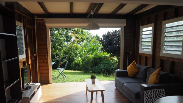 A modern living room with a sofa, a coffee table, and a TV. There is a sliding glass door leading to a garden with a chair and a table.