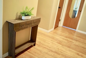 Una consola de madera con una planta en maceta y la inscripción 'Live Laugh Love', ubicada en un pasillo con piso de madera.